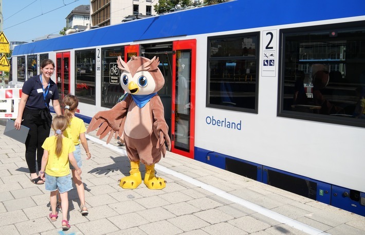 Das BRB-Maskottchen Loksi steht am Gleis begrüßt die Kinder zusammen mit einer Mitarbeiterin. Es sind 3 Kinder von hinten zu sehen.