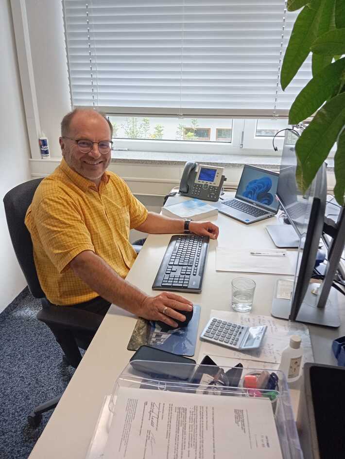 Unser Kollege Stefan Mühlstrasser sitzt an seinem Arbeitstisch an seinem PC und klacht in die Kamera.